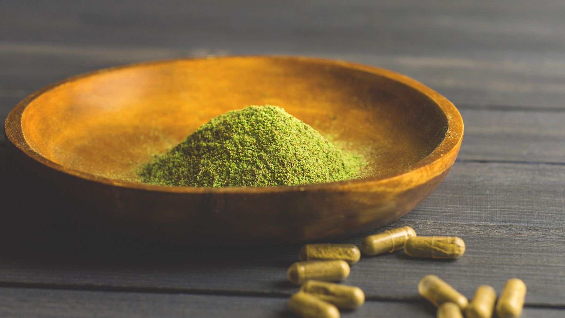 Green powder in wooden bowl with supplement capsules on dark surface